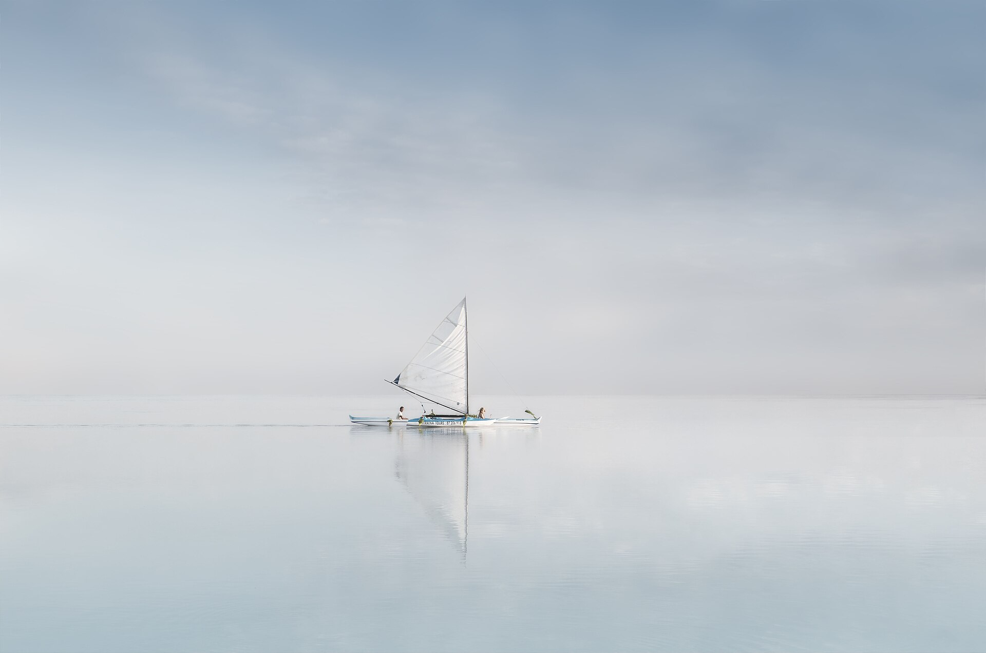 An outrigger canoe with sail on a glassy lagoon